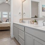A modern bathroom with gray cabinets, double sinks, and a large mirror, leading into a bright bedroom with a beige bed. Art hangs on the bathroom and bedroom walls, and natural light streams in through shuttered windows.