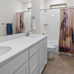 A clean bathroom with a double sink vanity, white cabinets, a toilet, and a shower with a curtain featuring a horse design. The room has neutral walls, tiled floor, and blue towels on silver towel racks.