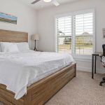 A bright bedroom with a wooden bed, white bedding, two paintings on the walls, a desk with an office chair, a lamp, and a large window with white shutters letting in natural light.