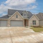 A modern two-story brick house with a three-car garage, sloped roof, and neatly landscaped front yard under a partly cloudy sky in a suburban neighborhood.