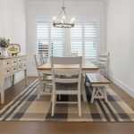A bright dining room with a wooden table, white chairs, a bench, a plaid rug, a buffet with decor on the left, and a chandelier hanging from the ceiling. Large shuttered windows let in natural light.