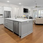 Modern open-concept kitchen and living area with white cabinets, stainless steel appliances, a large gray island, light wood floors, a beige sectional sofa, a wall-mounted TV, and large windows letting in natural light.