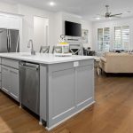 Modern open-concept kitchen and living room with a large gray island, white cabinetry, stainless steel appliances, hardwood floors, light-colored sofa, wall-mounted TV, and large windows with plantation shutters.