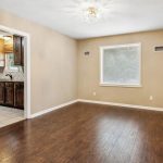 A bright, empty dining area with wood floors and beige walls connects to a kitchen with dark wood cabinets and tiled flooring; a window lets in natural light.