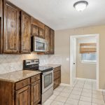 A kitchen with wooden cabinets, stainless steel appliances, granite countertops, and beige tiled floors. There is a window with a bamboo shade in the adjacent laundry or pantry area.
