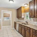 A kitchen with wooden cabinets, granite countertops, stainless steel appliances, a double sink under a window, and tiled floors and backsplash. An adjacent room with a window and blinds is visible through an open doorway.