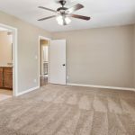 A spacious, empty bedroom with beige walls and carpet, a ceiling fan with lights, an open door leading to a bathroom with wooden cabinets, and three windows with white shutters letting in natural light.