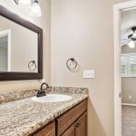 A bathroom with a granite countertop, white sink, wooden vanity, large mirror, and black fixtures. An open door leads to a carpeted bedroom with a ceiling fan and window shutters.