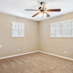 Empty beige room with carpeted floor, two windows with white shutters, a ceiling fan with lights, and visible wall scuff marks on the right.