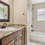 A bathroom with a granite countertop, wooden vanity, white sink, large framed mirror, and a bathtub-shower combo with beige tile walls and a window above the tub.