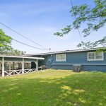 A blue single-story house with white trim features a covered patio area and sits on a grassy lawn with trees and an air conditioning unit visible near the house.