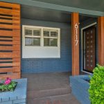 A modern house entrance with blue-gray brick, a dark door with glass squares, vertical wooden posts showing the address 6117, horizontal wooden slats, and a small flower bed with colorful plants in front.