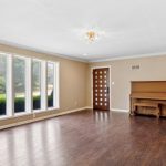 Spacious living room with light brown walls, dark wood flooring, three tall windows, a wooden front door with decorative glass, and a wooden upright piano against the far wall.