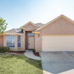 A single-story brick house with a two-car garage, large front windows, a light-colored front door, a small porch, and minimal landscaping with a tree and some bushes in the front yard.