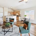 Bright, open-concept living and dining area with wood floors, green chairs, a dark sofa, and a round table. The kitchen has light wood cabinets, a breakfast bar with stools, and large windows let in natural light.