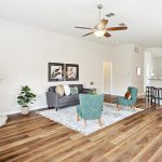Bright, open living room with wood flooring, modern furniture, a fireplace, ceiling fan, and a kitchen area with a dining table and chairs in the background. Plants and artwork add decorative touches.