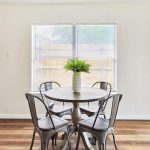 A round dining table with four metal chairs, a white vase with green ferns in the center, sits by a large window in a bright room with wooden floors and light-colored walls. A door to the outside is on the right.