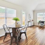 Bright dining and living area with large windows, wood flooring, a round table with four metal chairs, green armchair, small decor items, and views of a fenced backyard.