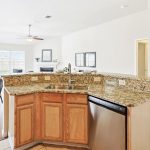 Modern open-concept kitchen with granite countertops, wooden cabinets, and stainless steel appliances. Kitchen overlooks dining area with metal chairs and living room with sofas and large windows.