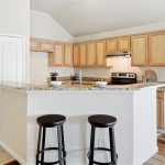 A modern kitchen with light wood cabinets, granite countertops, stainless steel appliances, and two black barstools at a breakfast bar. The space has wood and tile flooring and opens to a bright living area.