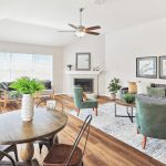 Bright living and dining room with wood floors, a round dining table, green chairs, gray sofa, potted plants, large windows, framed art, and a fireplace with a mirror above it.