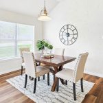 A bright dining area with a wooden table and four upholstered chairs on a patterned rug, large wall clock, decorative plants, and light wood flooring near a window letting in natural light.