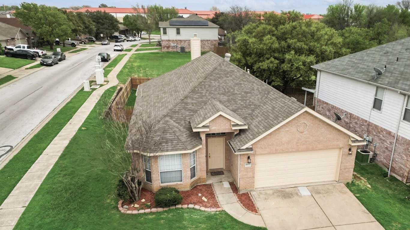 Aerial view of a single-story brick house with a two-car garage, neatly manicured lawn, and a curved walkway leading to the front door, located on a suburban street lined with trees and neighboring homes.