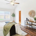 A bright bedroom with a neatly made bed, decorative pillows, a green throw, wooden flooring, a ceiling fan, a window with outdoor view, and a console table with plants and decor against a white wall.
