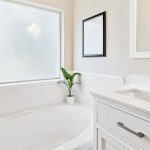 Bright bathroom with a white bathtub, a frosted window, a small potted plant on the tub ledge, a white vanity with a sink, and a framed picture on the wall.