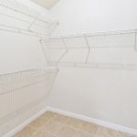 Empty walk-in closet with white wire shelving on three walls, beige tiled floor, and white textured walls. The shelving is arranged at different heights for storage.
