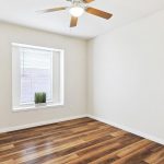A small, empty room with white walls, wood-look flooring, a window with blinds, a potted plant on the windowsill, and a ceiling fan with a light.