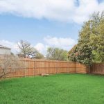 A backyard with green grass, a wooden fence, a small tree, and several large trees along the fence. There is a house partially visible on the left and another building just beyond the fence. The sky is partly cloudy.