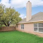A fenced backyard with green grass, a large tree, and a brick house featuring several windows and a tall chimney under a partly cloudy sky.
