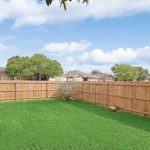 A fenced backyard with bright green grass, a small shrub near the fence corner, and houses and trees visible beyond the wooden fence under a partly cloudy sky.
