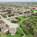 Aerial view of a suburban neighborhood with houses, tree-lined streets, green spaces, and a nearby large building, possibly a school, surrounded by a forested area and open fields.