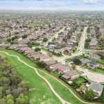 Aerial view of a suburban neighborhood with rows of houses, curved streets, and green spaces bordered by trees under a cloudy sky.