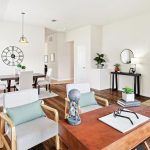A bright, modern open-concept living space with a desk and chairs in the foreground, a dining table and chairs in the background, plants, wall decor, and wood floors throughout.