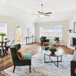 Bright, modern living room with wood floors, a gray sofa, green chairs, a round coffee table, a dining area with metal chairs, large windows, white walls, and a corner fireplace with decorative accents.