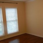 An empty room with light beige walls, two large windows with white blinds, a curtain rod above, and a hardwood floor. There are electrical outlets on the right wall.