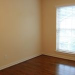 Empty room with beige walls, two large windows with white blinds on the right, and a dark wood floor. Natural light is coming through the windows, illuminating the space.