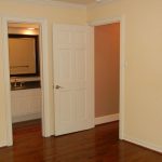 An empty room with beige walls and dark wooden floors. One door is open, revealing a bathroom with a sink and mirror, while a second doorway leads to another room.
