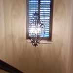 An ornate metal chandelier hangs in a stairwell, lit with several bulbs. Behind it is a window with wooden shutters, casting shadows on the cream-colored walls. A dark wooden handrail is visible on the left.