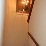 A staircase with wooden handrails leads up to a landing with a window featuring wooden shutters. A decorative hanging light fixture is illuminated above the landing, casting shadows on the wall.