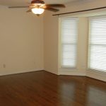 Empty room with light beige walls, dark wood flooring, a ceiling fan with a light, and a large bay window with white blinds letting in natural light.