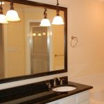 A bathroom with a large mirror above a black granite countertop with two sinks, three pendant lights, a bathtub, white cabinets, and beige walls.