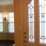 A decorative glass front door is partially open, revealing a living room with beige walls, white trim, dark wood floors, and large windows with blinds and a wall sconce.
