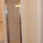 A bathroom with a glass shower door, a white bathtub with a metal faucet, beige walls, a toilet, and white cabinets on the right side.