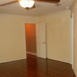 Empty room with beige walls, white trim, and wooden floor. A ceiling fan with a light is mounted above. An open white door leads to a hallway with an orange wall visible.
