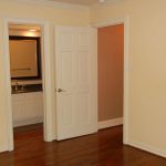 Empty room with light beige walls and wooden floor. One door is open, showing a bathroom with a sink and mirror, while another door opens to a hallway. Room has simple white trim and a wall outlet.