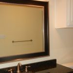 A bathroom with a large framed mirror above a black granite countertop and sink, a towel bar on the opposite wall, and a shower with beige tiles and a white cabinet above the toilet.